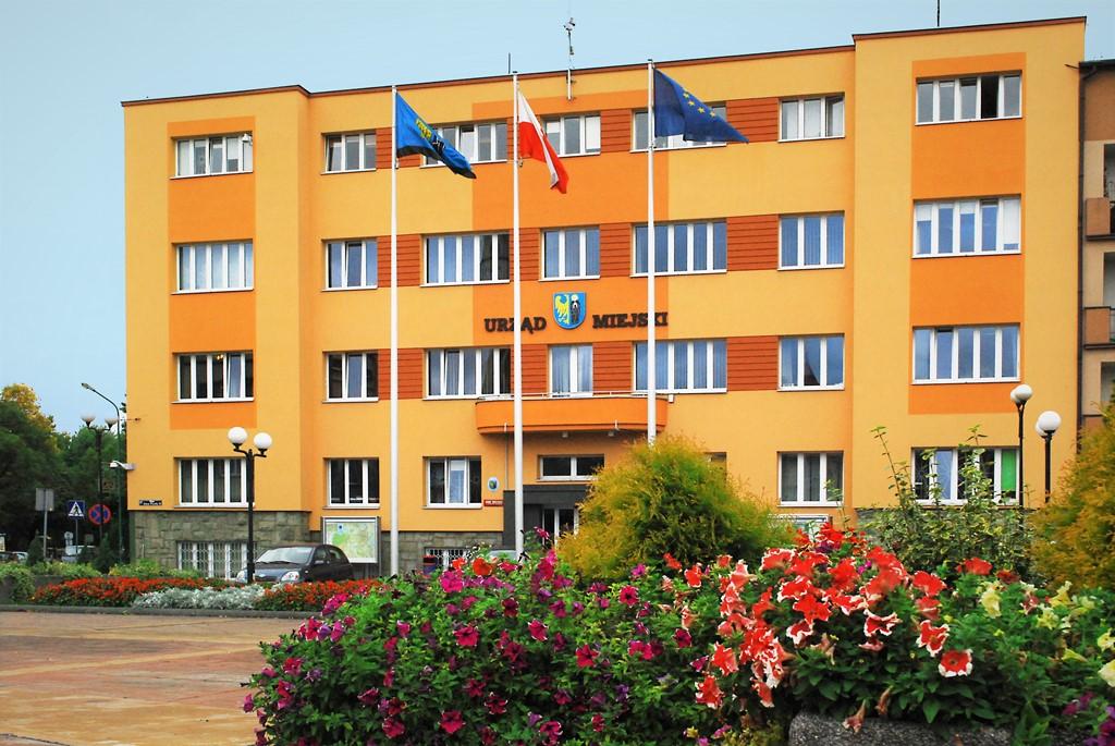 w tle bydynek urzędu miejskiego w Czechowicach-Dziedzicach, przed nim widoczne krzewy oraz flagi: miejska, polska i unijna