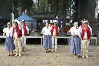 pierwszy plan: przed sceną stoją tancerze regionalnego zespołu, dziewczyny są ubrane w białe bluski i granatowe spódnice, chłopcy mają czerwone kamizelki, na głowie kapelusze, stoją parami, w tle scena