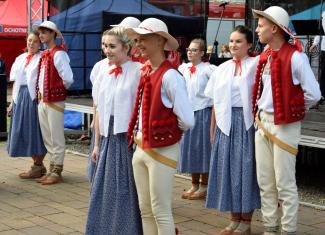 przed sceną stoją tancerze regionalnego zespołu, dziewczyny są ubrane w białe bluski i granatowe spódnice, chłopcy mają czerwone kamizelki, na głowie kapelusze, stoją parami, w tle scena