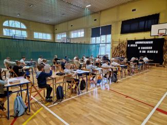 dzieci siedzące przy stolikach ustawionych na sali gimnastycznej, w trakcie pisania konkursu